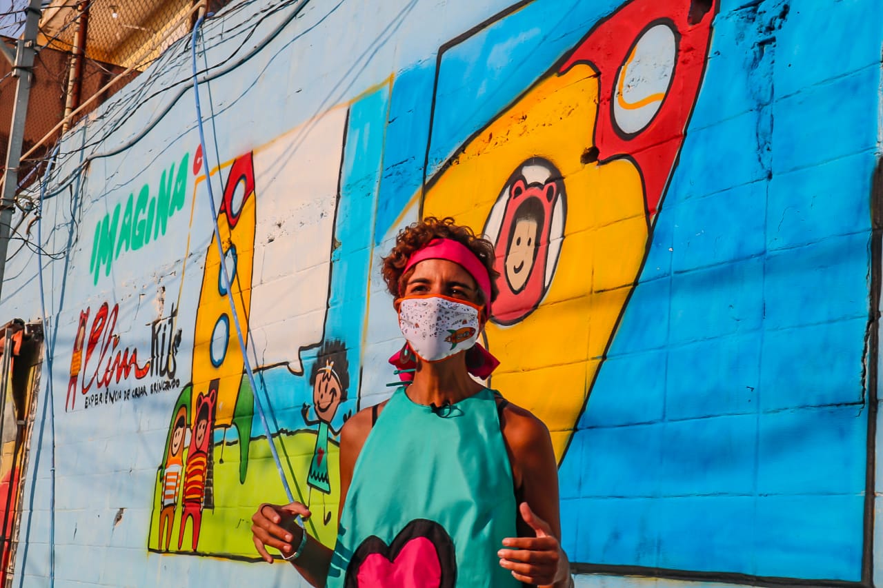 Mulher com vestido verde com coração rosa, faixa rosa e máscara e muro ao fundo pintado e colorido 